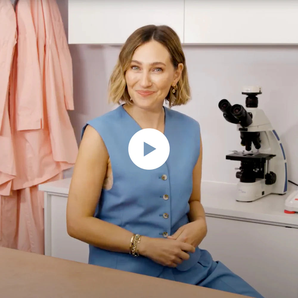 A woman with short, wavy blonde hair in a blue sleeveless vest sits in a lab beside a microscope and pink lab coats, smiling at the camera. There is a play button over the image, indicating a video.