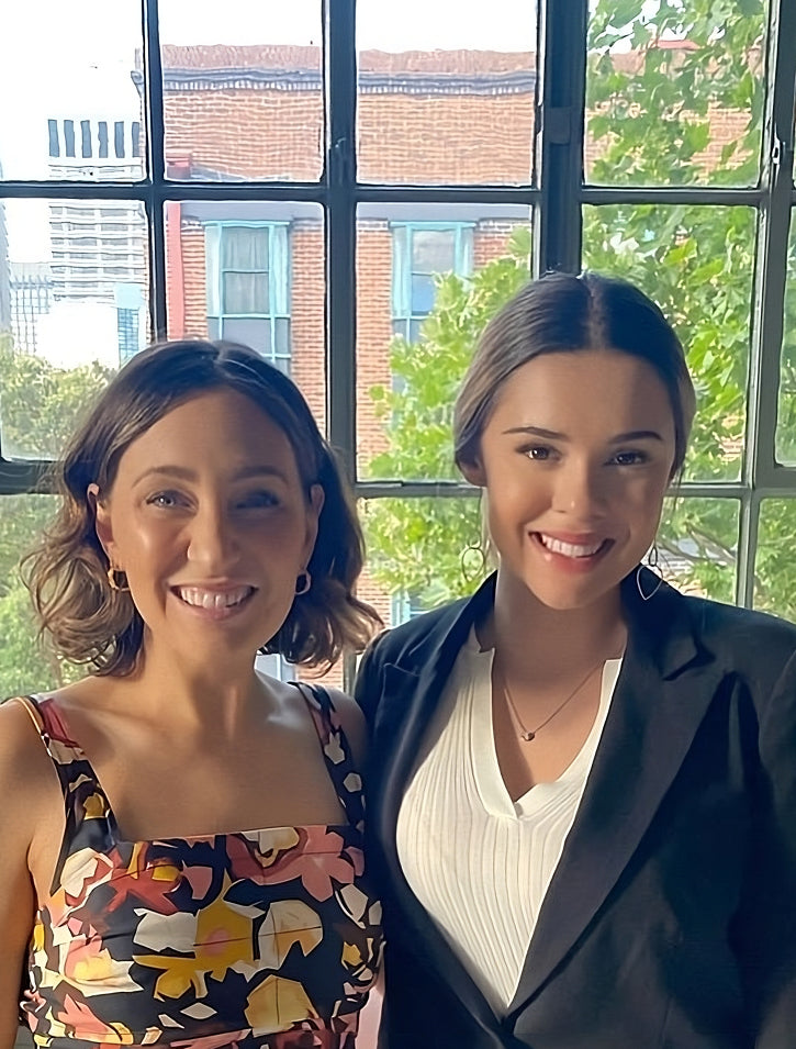 Two women smiling indoors in front of large windows. One wears a colorful floral dress, the other a black blazer over a white top. Green trees and brick buildings are visible outside through the windows.