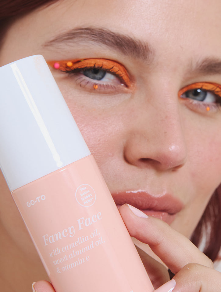 A woman with orange eye makeup and decorative gems holds a peach-colored bottle of Fancy Face skincare product with a white cap close to her face. The label mentions camellia oil, sweet almond oil, and vitamin E.