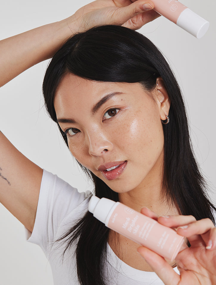 A woman with long black hair holds two pink skincare product bottles, one near her face and one above her head, while posing with glowing, dewy skin and wearing a white shirt against a plain background.