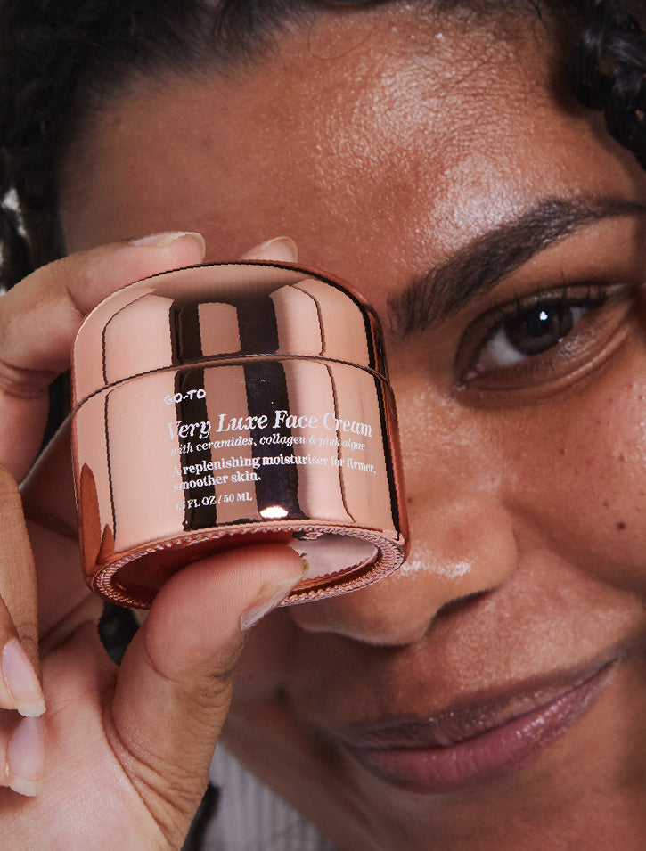 A person with curly hair holds a small, rose gold jar labeled Very Luxe Face Cream close to their face, smiling softly and looking into the camera.