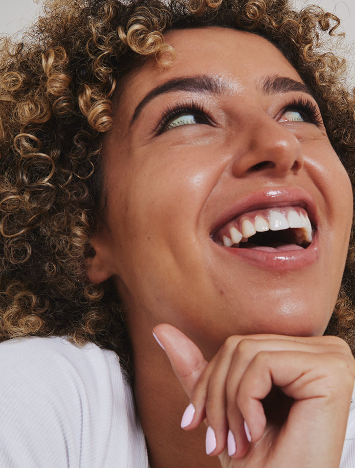 A person with curly hair smiles brightly, showing their teeth, while looking upward. Their hand is near their chin and they wear light pink nail polish. The image captures a joyful and expressive moment.