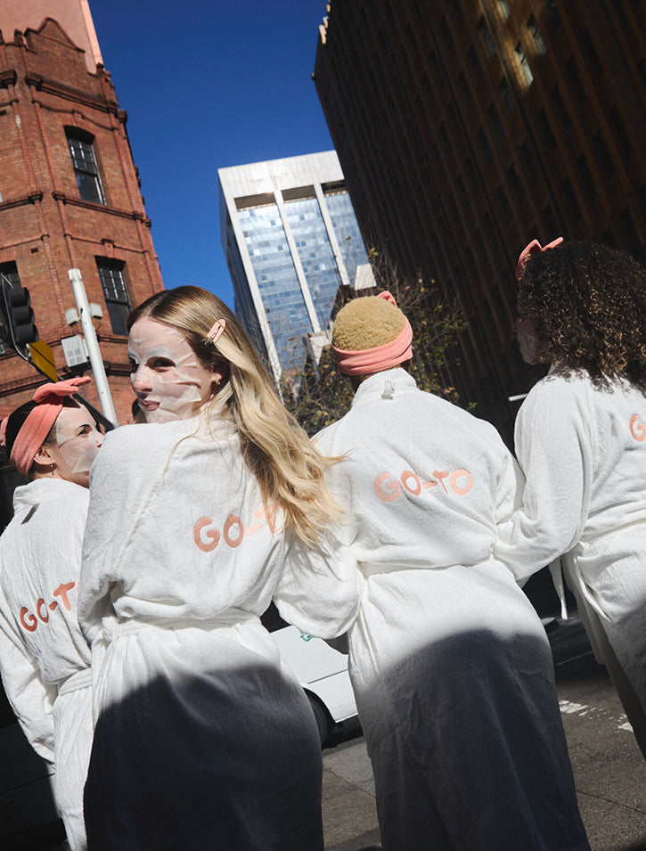 Four people wearing white robes and sheet face masks cross a city street. The robes say GO-TO on the back. Tall buildings and a clear blue sky are visible in the background.