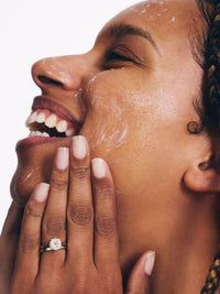 A woman with braided hair smiles, applying skincare, her hand adorned with a sparkling ring, highlighting her radiant complexion.