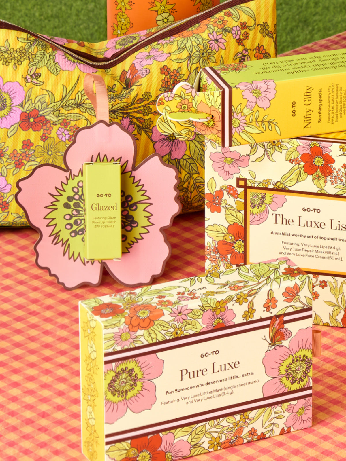 Go-To Skincare Glazed tinted SPF lip oil displayed with colorful floral gift boxes and a pink flower shaped paper holder on a pink checked surface