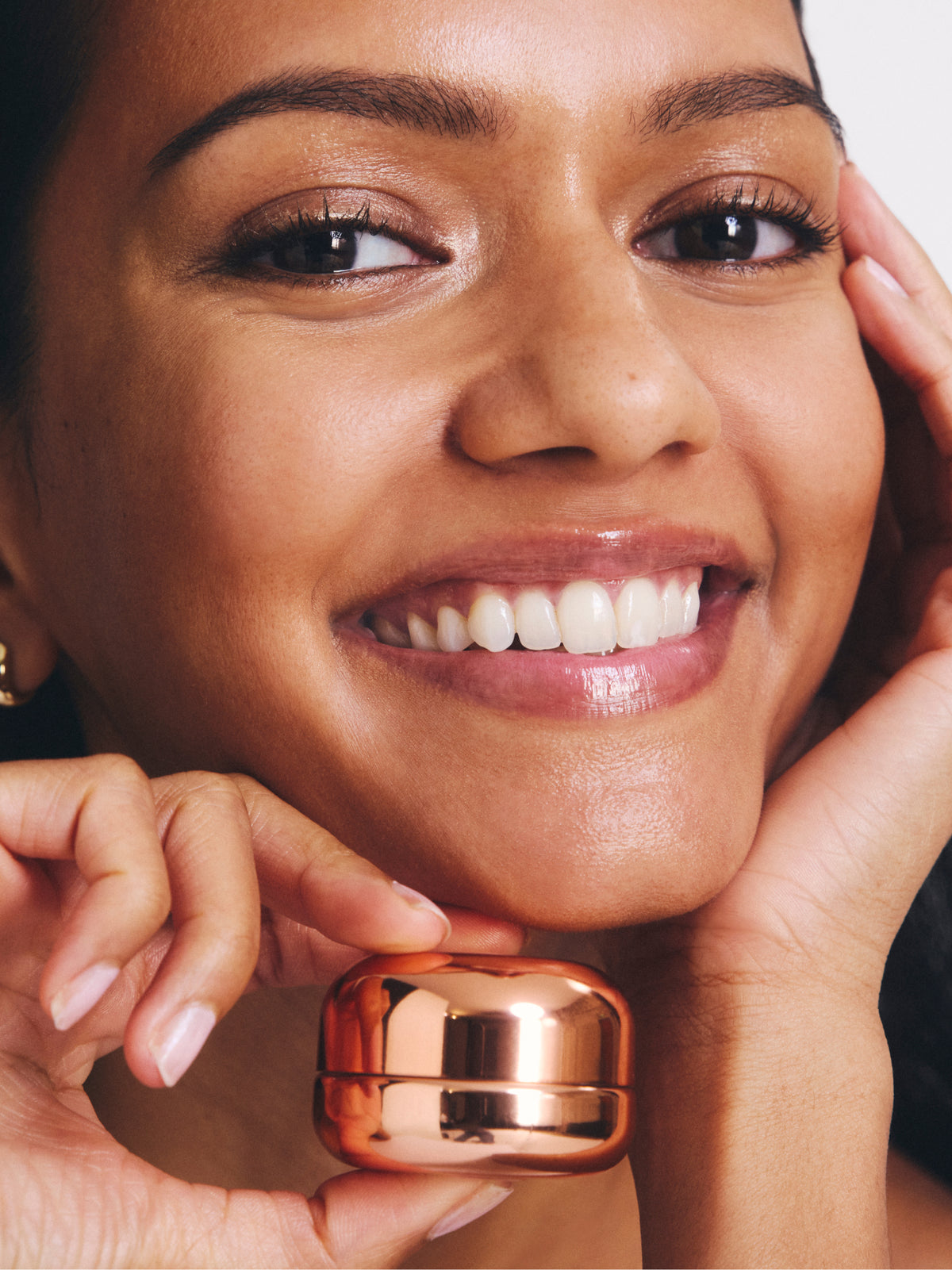 Close up of a smiling woman with radiant skin holding a reflective rose gold skincare container near her chin from Go To Skincare