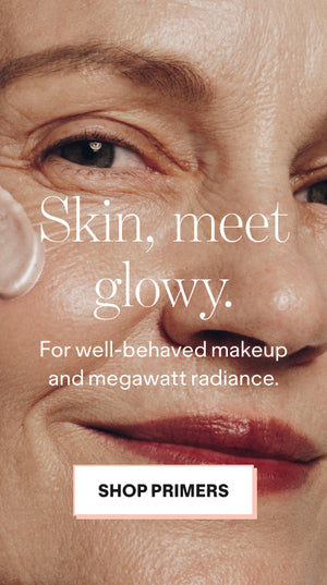 Close-up of a mature woman applying primer to her cheek, smiling softly. Overlaid text reads, Skin, meet glowy. For well-behaved makeup and megawatt radiance. A button below says, Shop Primers.