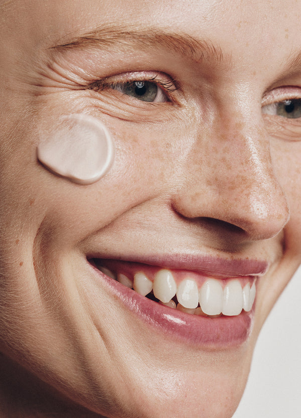 Close-up of a smiling person with light skin, blue eyes, and freckles, showing white cream applied to their cheek. Their teeth are visible and their skin appears smooth and healthy.