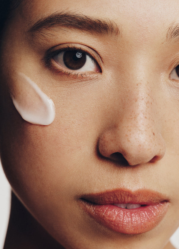 Close-up of a womans face showing natural skin texture, freckles, and a small swipe of white cream on her cheek. Her lips are slightly parted and she has a calm expression.