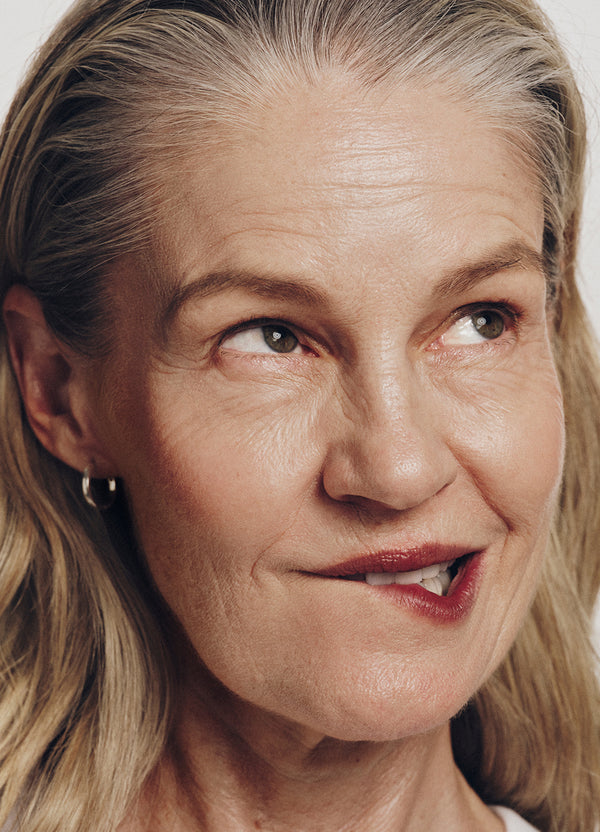 A close-up of a middle-aged woman with light skin and long, blondish-gray hair, looking slightly to the side and biting her lower lip with a playful expression. She is wearing a small hoop earring.