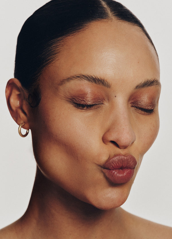A person with smooth skin and subtle bronze eye makeup closes their eyes and puckers their lips in a kiss, wearing a gold hoop earring and their dark hair pulled back.