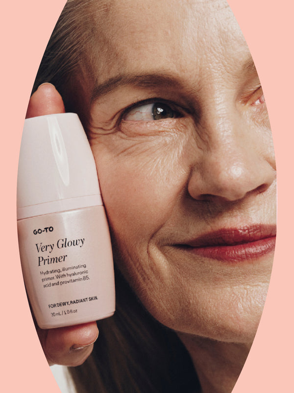 A smiling older woman holds up a pink bottle of Go-To Very Glowy Primer next to her face. The background is a soft pink, and the primer label highlights hydrating and illuminating properties for dewy, radiant skin.