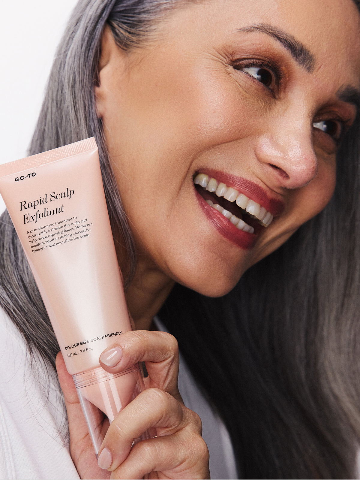Smiling woman with long dark hair holding a pink Go-To Skincare Rapid Scalp Exfoliant tube beside her hair