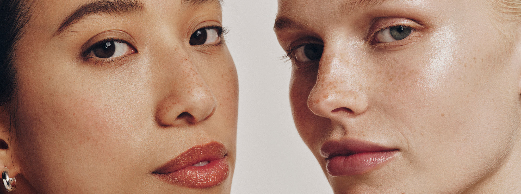 Close-up of two women with natural makeup and glowing skin, showing freckles and smooth complexions, set against a neutral background.
