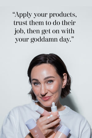 A smiling woman with short brown hair holds several skincare products, standing in front of a wall with the text: Apply your products, trust them to do their job, then get on with your goddamn day.