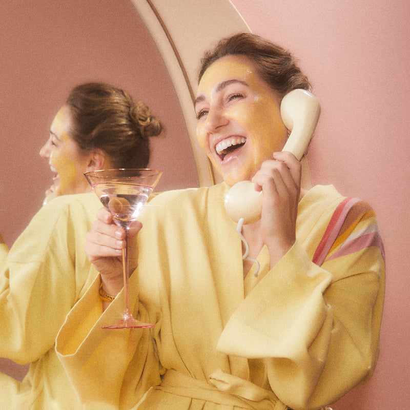 A smiling woman in a yellow robe holds a martini glass and talks on a vintage phone. She has a yellow face mask on and stands in front of a mirror, with her reflection visible. The background is pink.