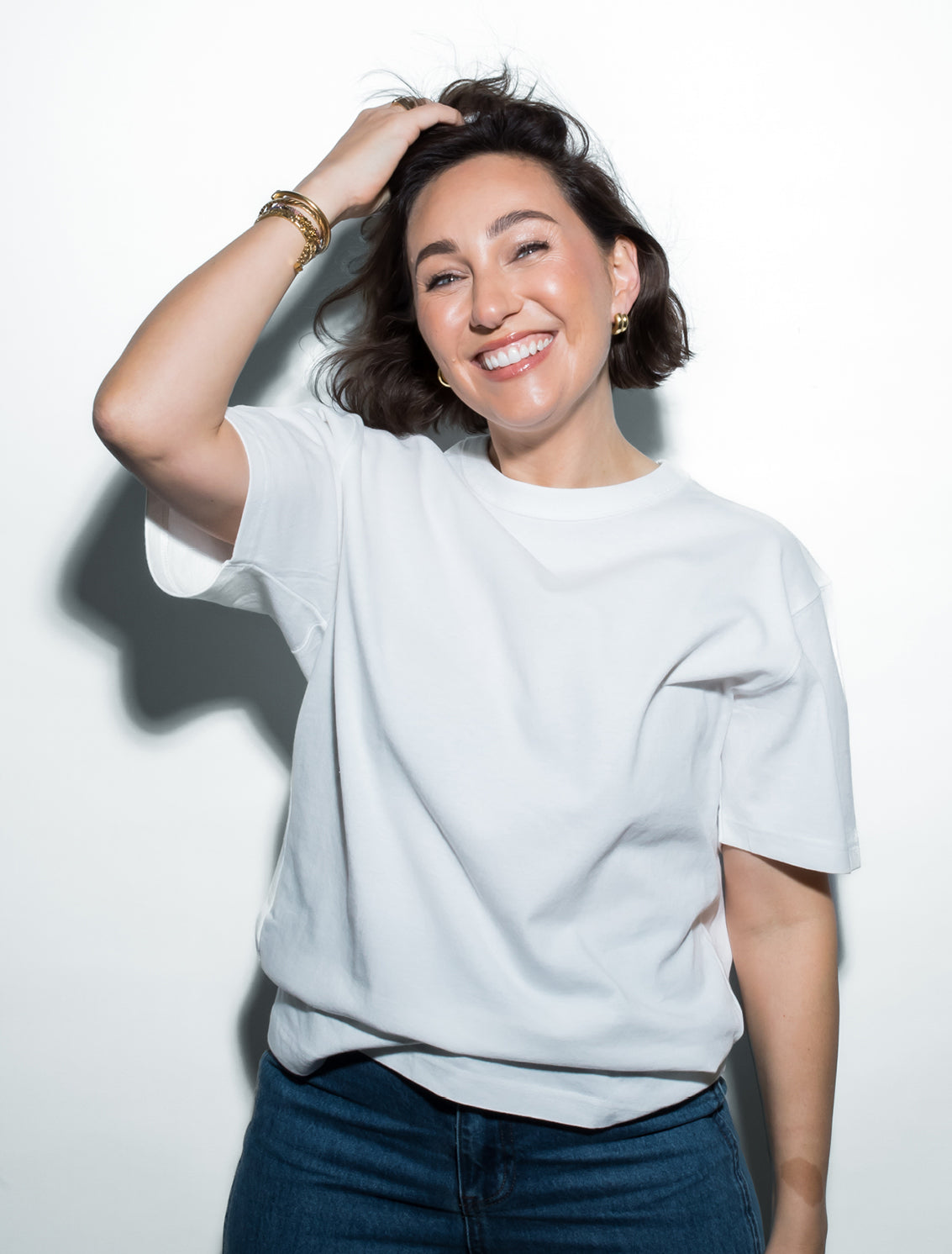 A woman with short dark hair smiles brightly, touching her head with one hand. She wears a white t-shirt, gold hoop earrings, bracelets, and blue jeans, standing against a plain white background.