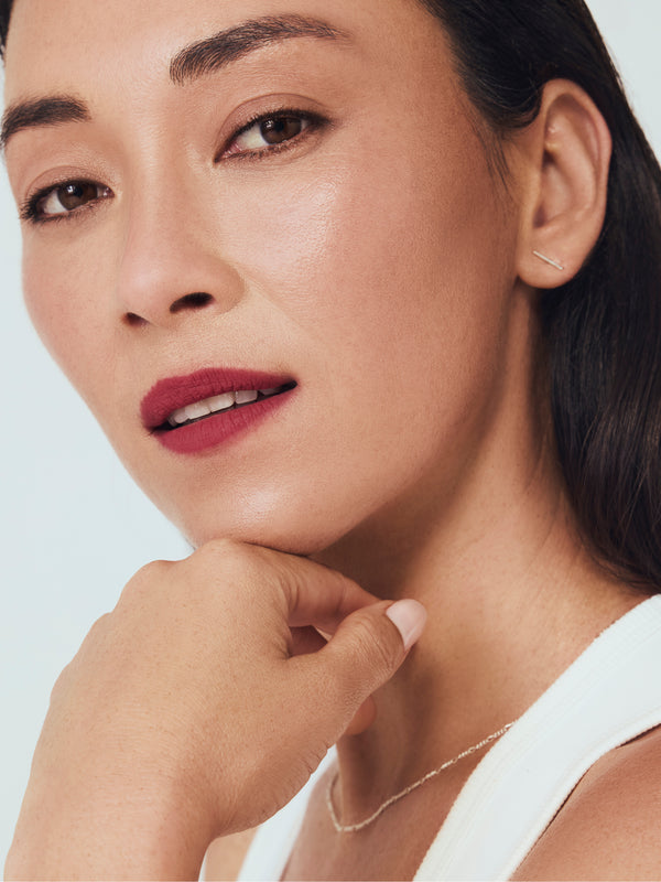 Woman with red lipstick and delicate jewelry poses thoughtfully, resting chin on hand against a soft background.