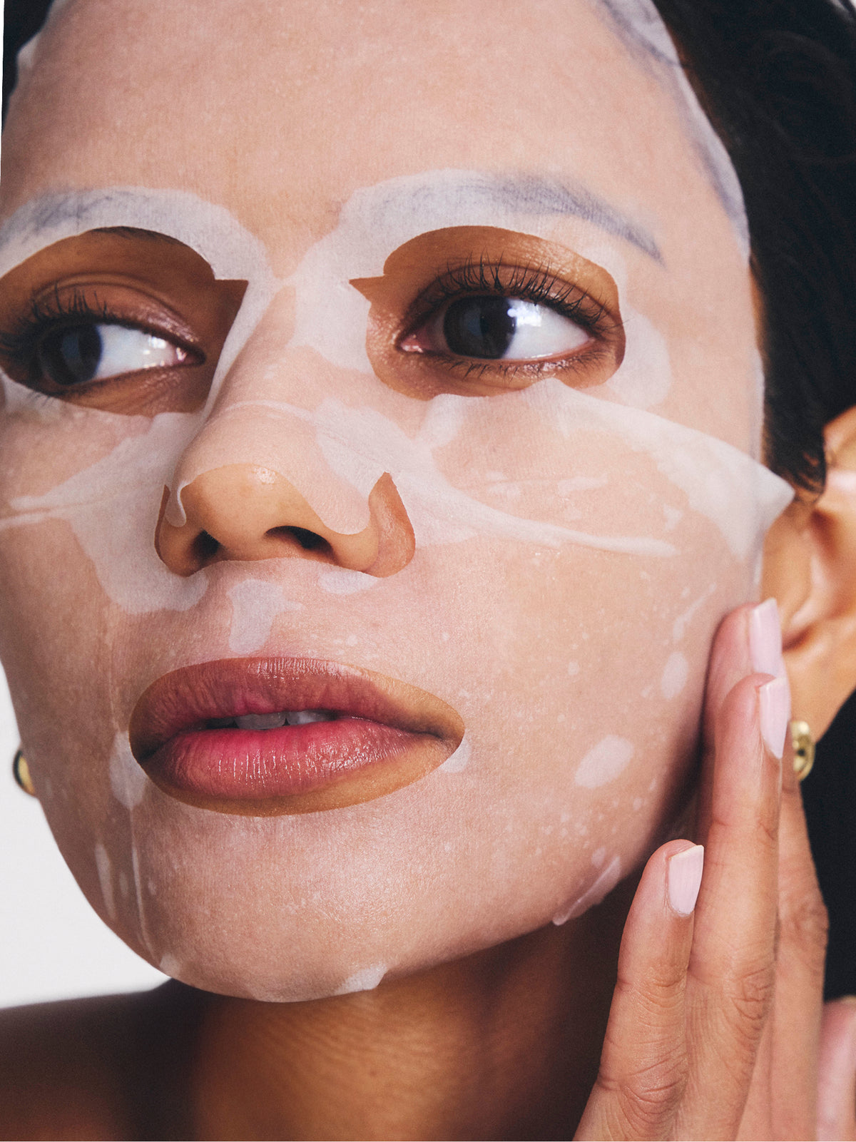 Close up of a woman wearing a white lifting sheet mask, with her hand gently touching her cheek and glossy lips visible.
