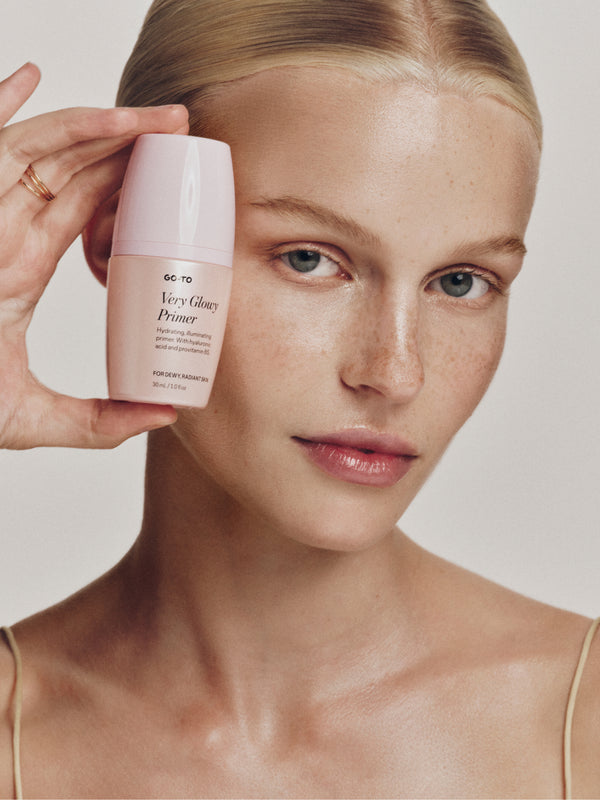 A person holds a Go-To Very Glowy Primer bottle near their face, showcasing smooth, radiant skin and a natural makeup look.