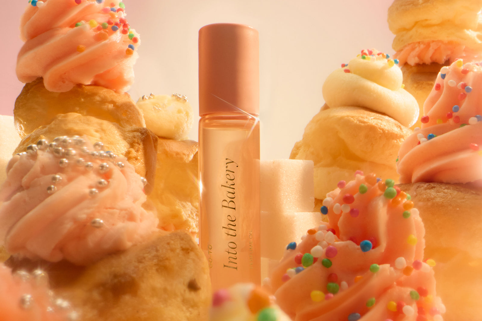 A perfume bottle labeled Into the Bakery is surrounded by cupcakes with pink frosting, cream puffs, colorful sprinkles, and sugar cubes, creating a sweet and whimsical bakery scene.