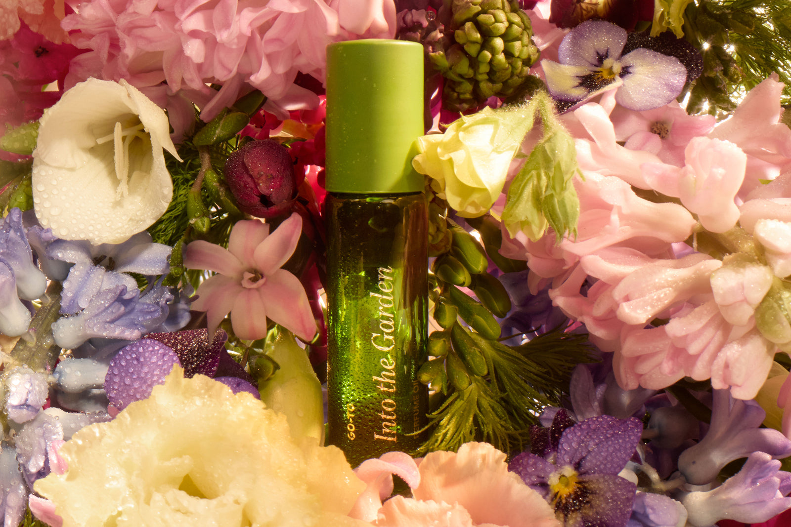 A green perfume bottle labeled Into the Garden is nestled among colorful, dewy flowers in shades of pink, purple, white, and yellow. The lush arrangement surrounds and highlights the bottle.