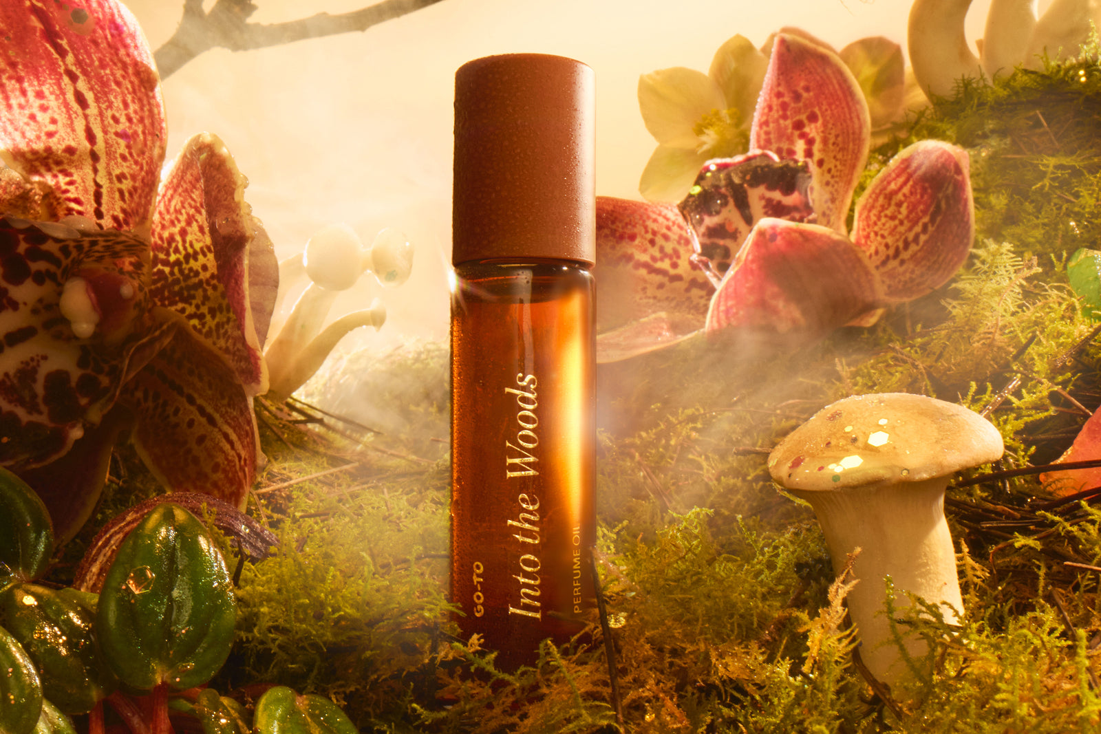 A brown glass perfume bottle labeled Into the Woods stands upright on moss, surrounded by mushrooms, green leaves, and pink orchids, with soft mist and warm light creating a dreamy, forest-like atmosphere.
