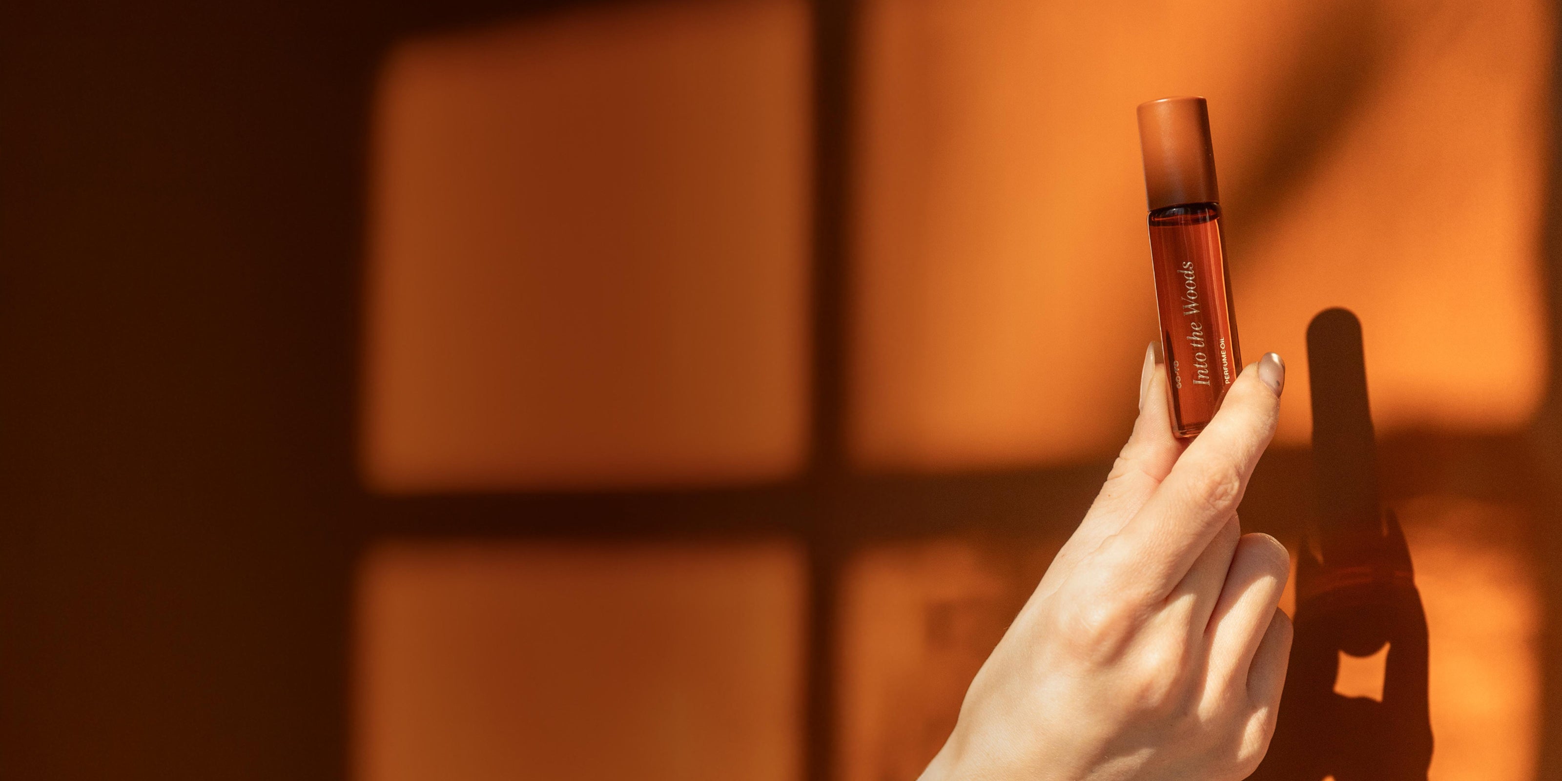 A hand holding a small brown bottle of perfume against a warm, orange background with soft shadows and window light.