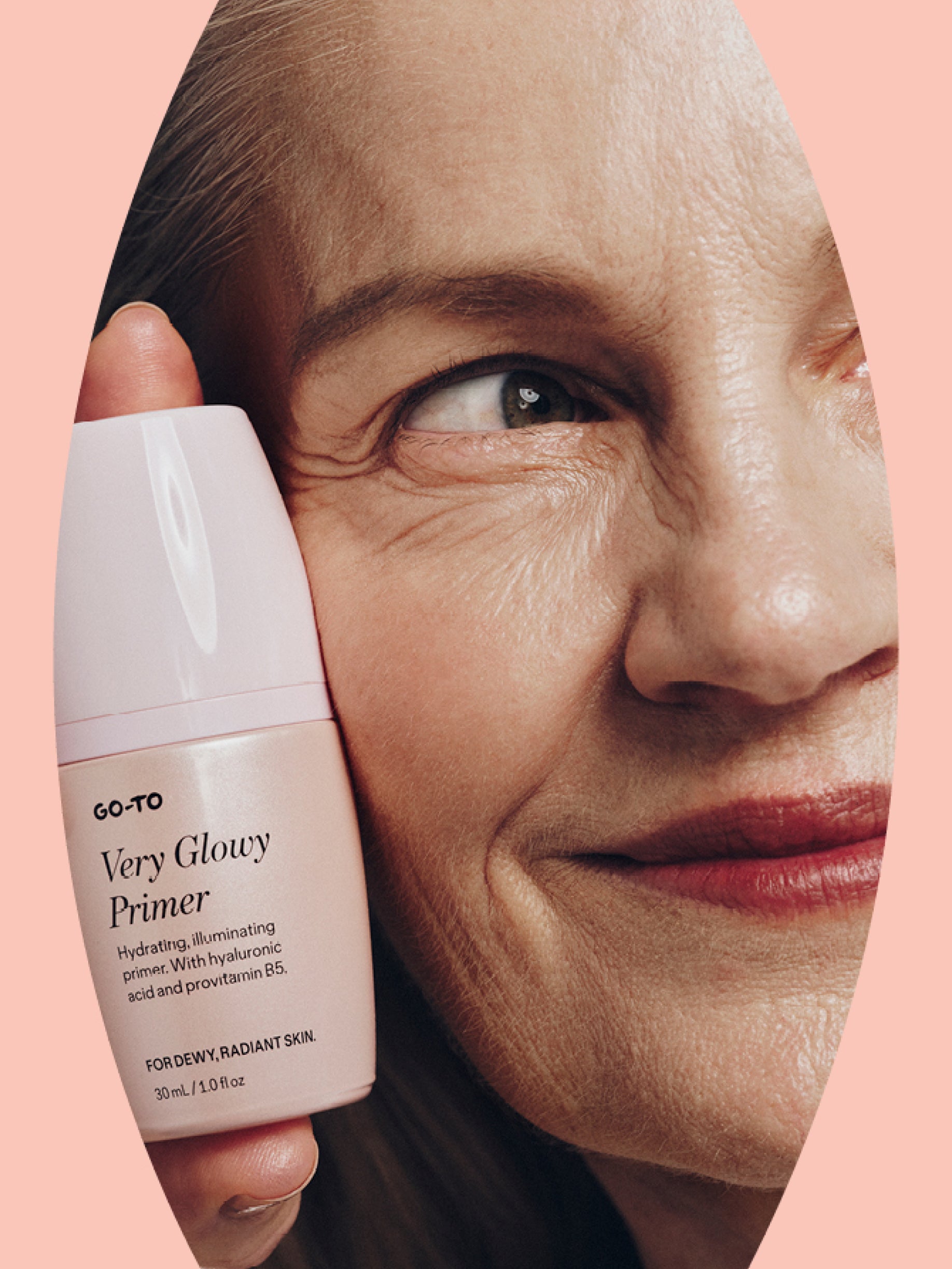 A smiling older woman with smooth skin holds a pink bottle of Go-To Very Glowy Primer close to her face, highlighting glowing, healthy skin against a peach background.