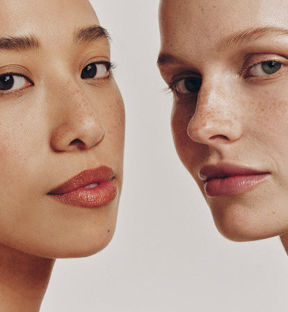Close-up of two women with natural makeup and smooth skin, standing side by side against a light background. One has dark hair and warm-toned skin; the other has light hair and fair skin. Both have a neutral facial expression.